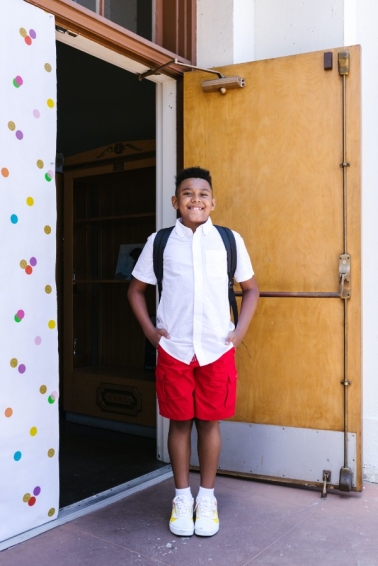 small boy smiling with hands in pockets standing outside a door